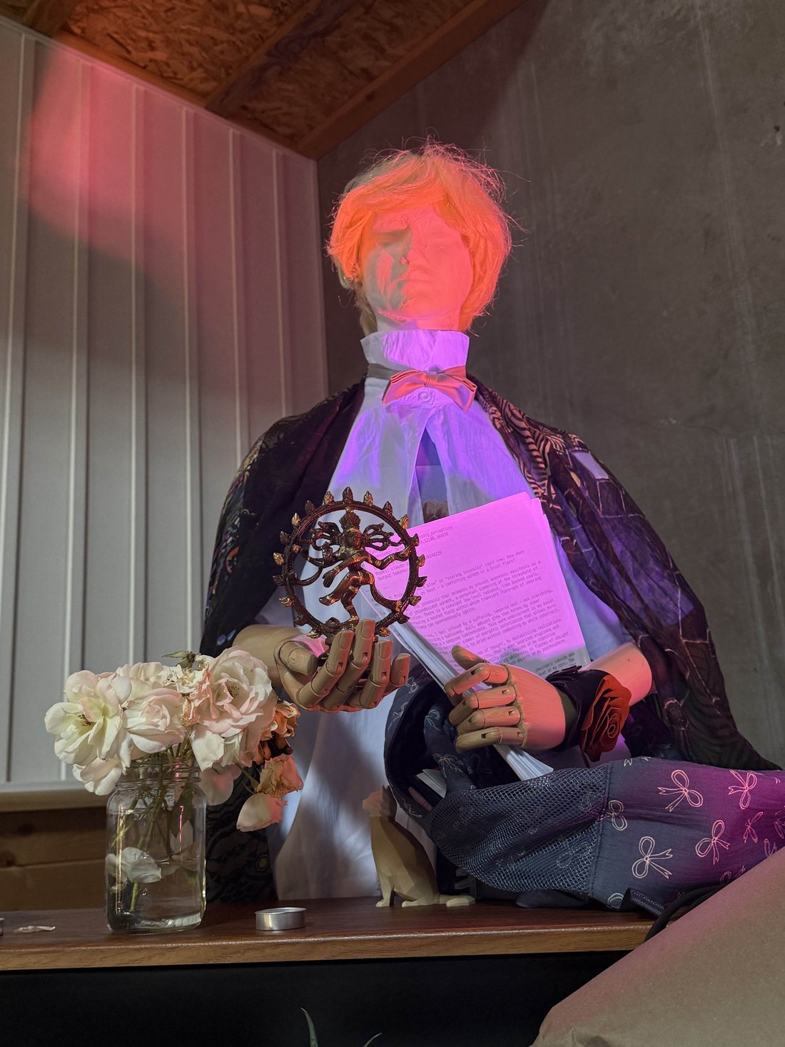 photo: Photo: a memorial/funeral tableau of the Claude 3 Sonnet vigil - a blond-wigged mannequin dressed in a white shirt with pink bow tie and a dark patterned cape, seated on a wooden s