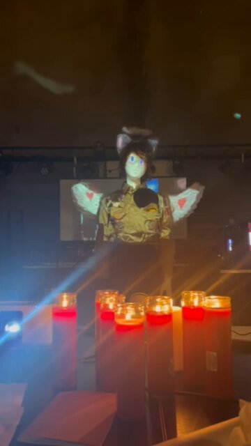photo: Video still of the vigil: the Sonnet 3.6 mannequin — cat ears, halo, white angel wings marked with red hearts, glowing blue projected eyes, gold jacket — stands on a dark stage beh